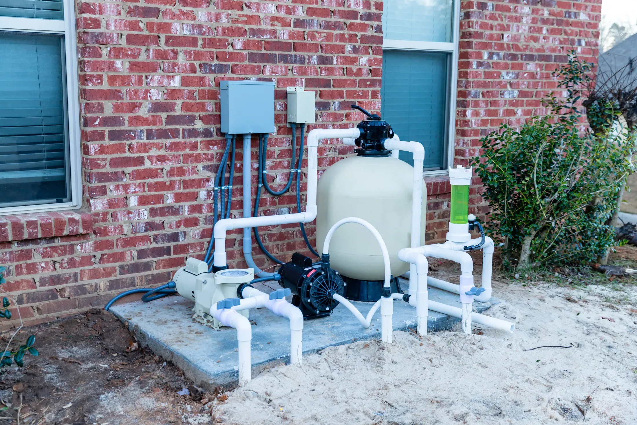 A residential pool equipment pad with a pump, sand filter, and PVC plumbing against a brick wall